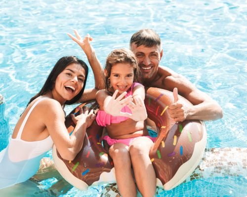 Excited young family having fun together at the swimming pool outdoors in summer, swimming with inflatable ring donut